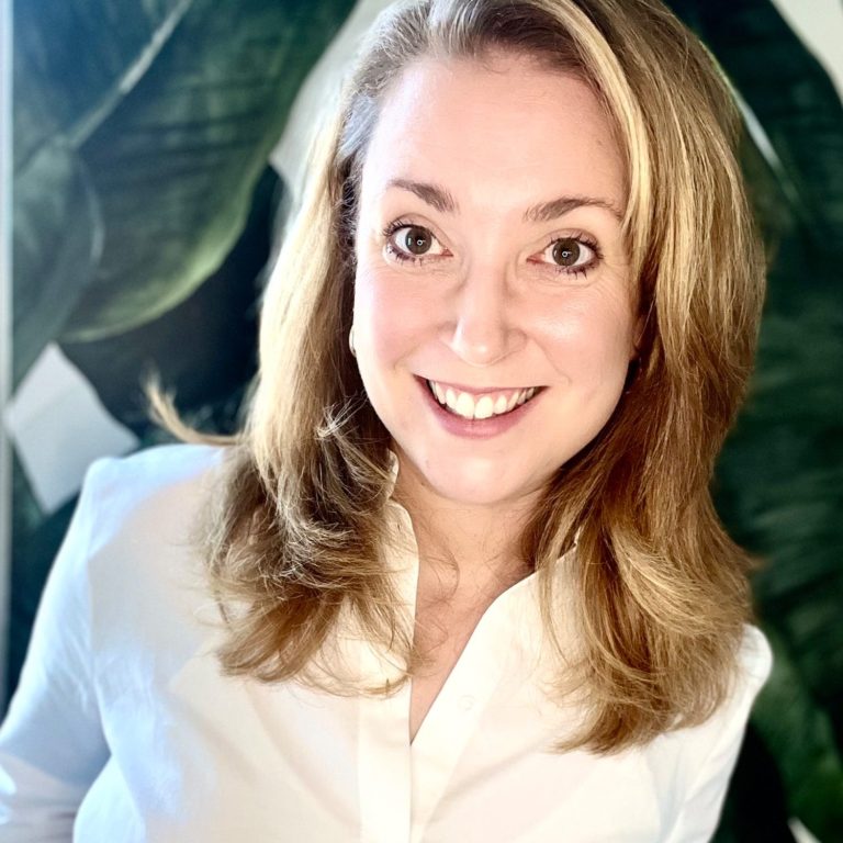 Katharina Henger Eine lächelnde Frau mit langen, hellblonden Haaren vor einem grünen Blatt Hintergrund.